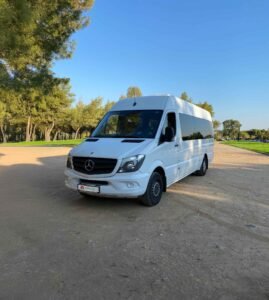 White Mercedes minibus parked on dirt road with pine trees and greenery under clear blue sky