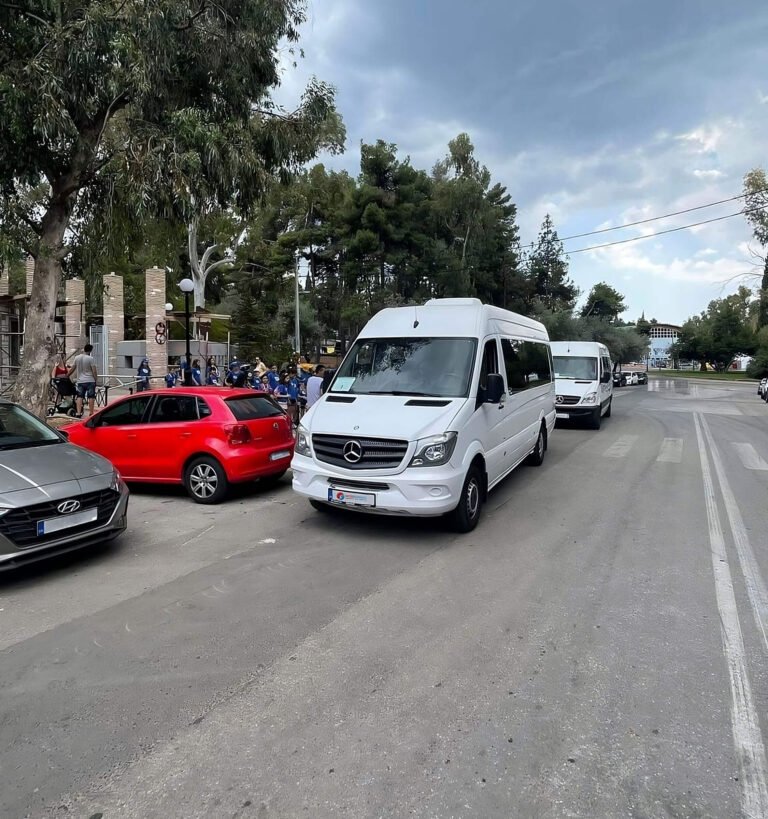 White Mercedes minibuses parked near a gate in an urban area with people and cars nearby